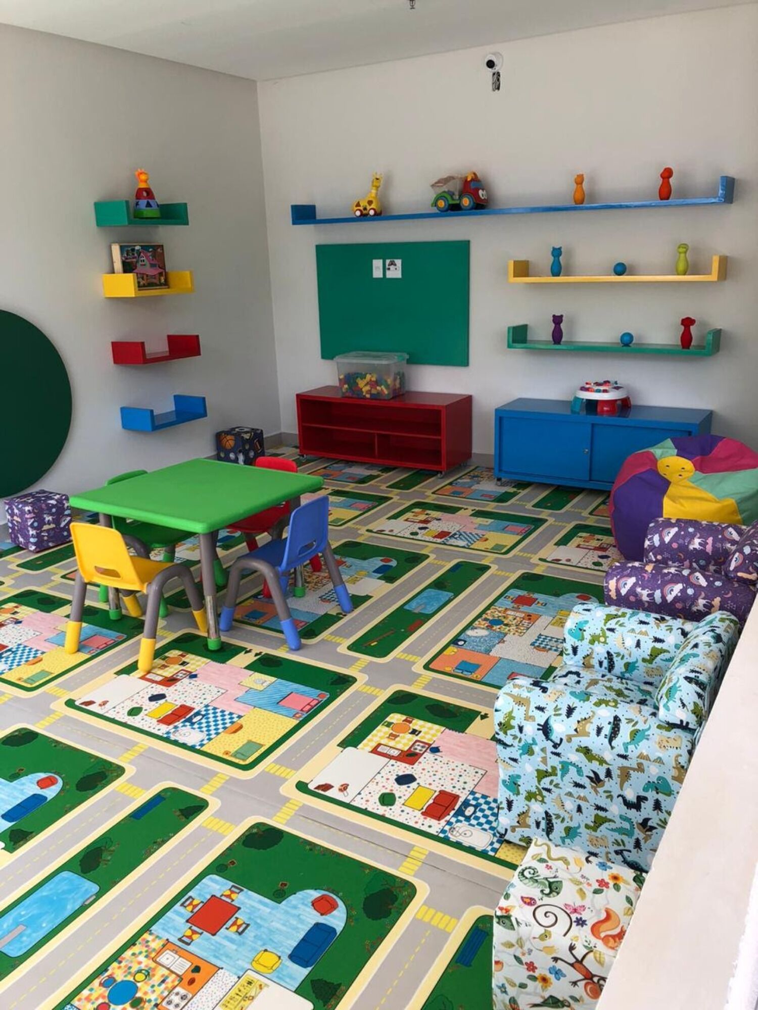 A children's playroom with colorful furniture, a green play mat, and educational toys on shelves.