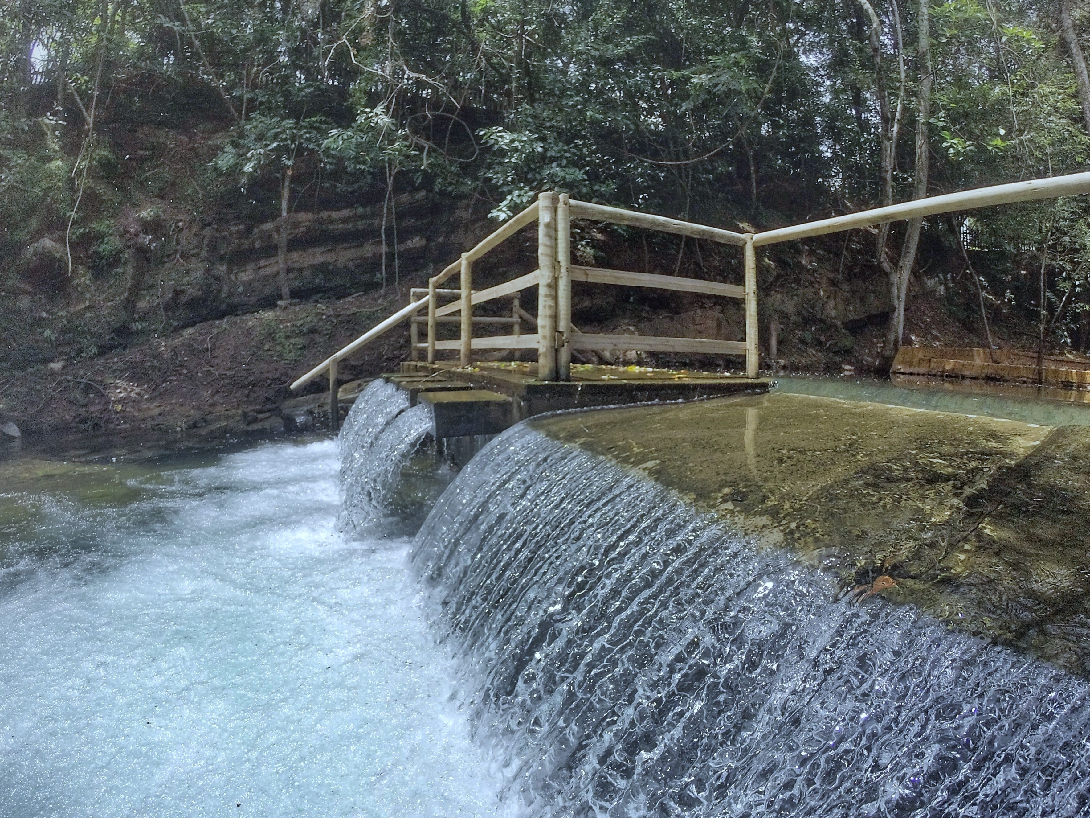 Eine Holzbrücke über einem Wasserfall mit steinigem Flussufer und dichtem Wald im Hintergrund.