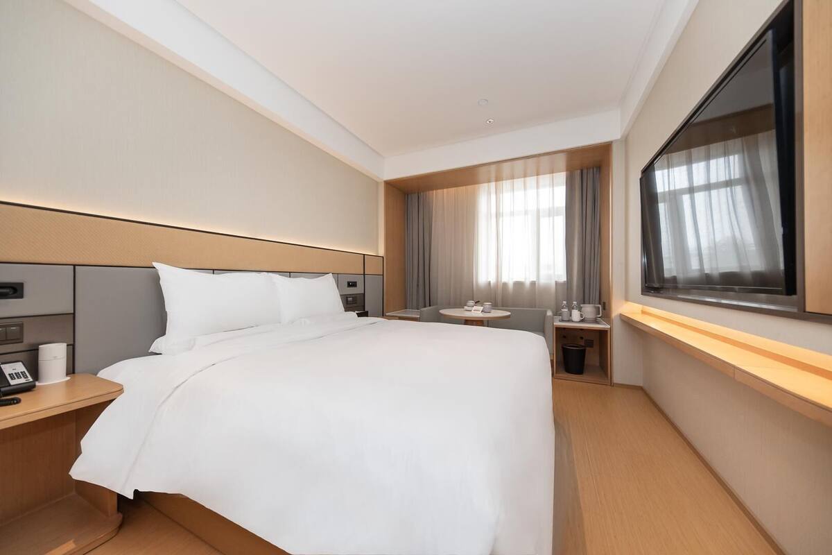 A hotel room with a large bed, bedside tables, a small table with a chair, a window with curtains, and a TV mounted on the wall.