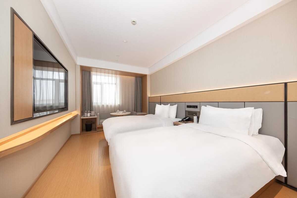 A hotel room with two beds, a desk, a chair, a large mirror, and a window with curtains.