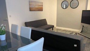 A modern living room with a glass dining table, grey sofa, and a flat-screen TV.