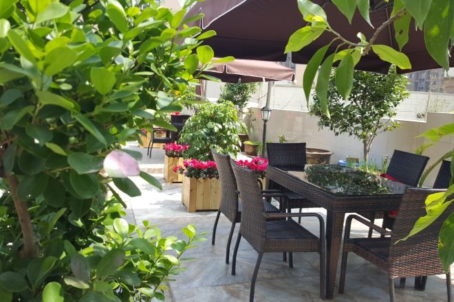 Un patio al aire libre con mobiliario de mimbre, plantas en macetas y una mesa bajo una zona sombreada.
