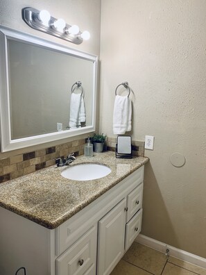 A bathroom with a granite countertop sink, a mirror with lights, a towel rack, and a potted plant.