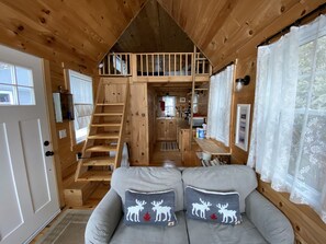 Interior de una cabaña con una escalera de madera que sube a un altillo, un sofá con almohadones de ciervo y una zona de cocina.