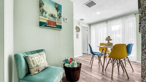 A living room with a teal sofa, a painting of a beach scene, and a dining area with yellow and blue chairs.