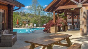 Ein hölzerner Picknicktisch in einem überdachten Außenbereich mit Pool und Liegestühlen im Hintergrund.