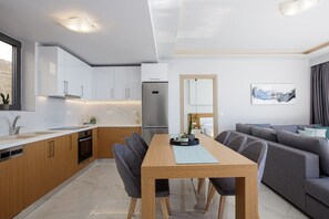 A modern kitchen with white cabinets, a built-in oven, and a stainless steel refrigerator. There is a dining table with chairs and a grey sofa in the living area.