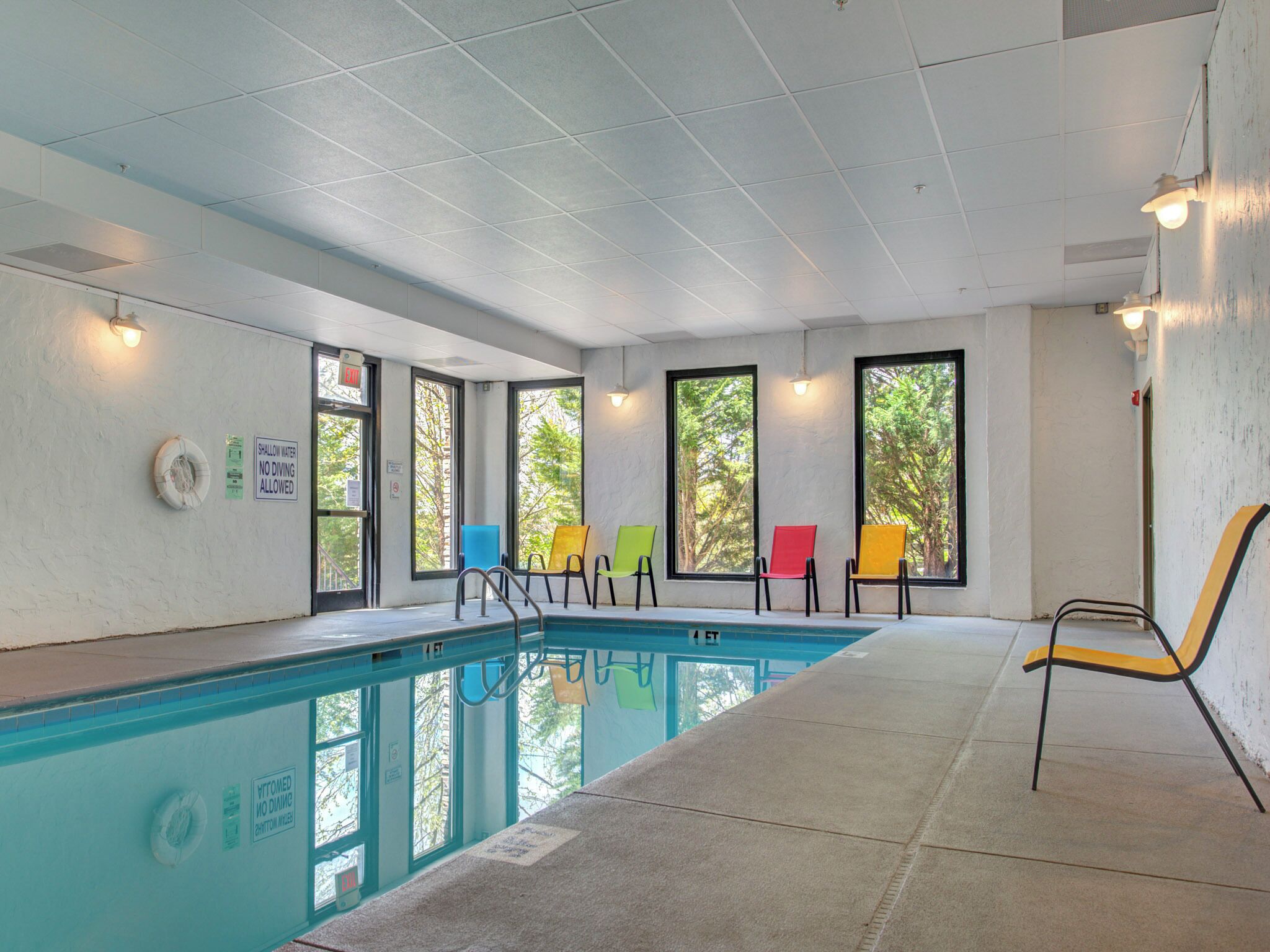 Una piscina con suelo de baldosas, ventanas con vistas a árboles y sillas coloridas a lo largo de la piscina.