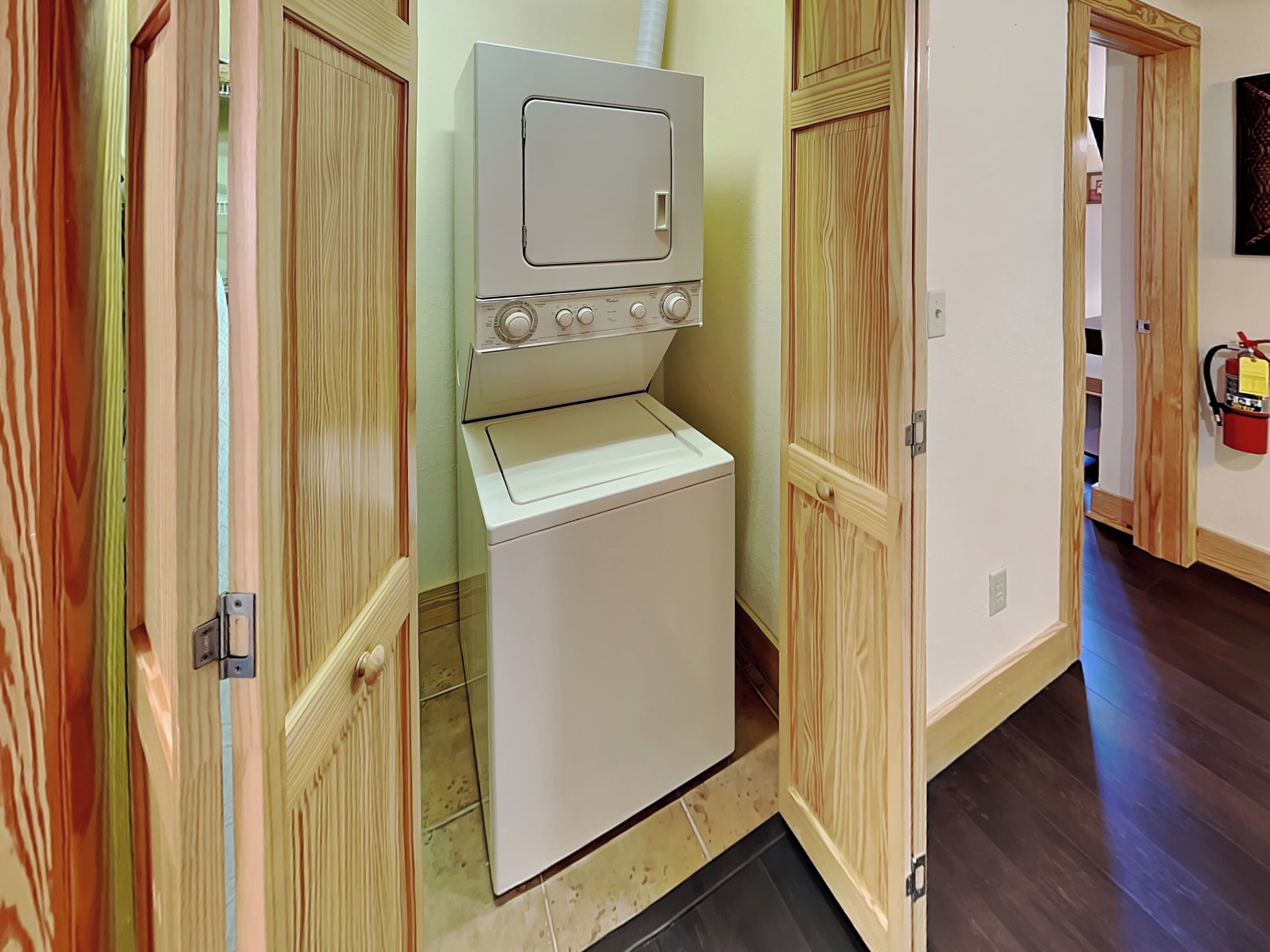 A compact laundry room with a front-loading washer and dryer, wooden doors, and a visible fire extinguisher on the wall.