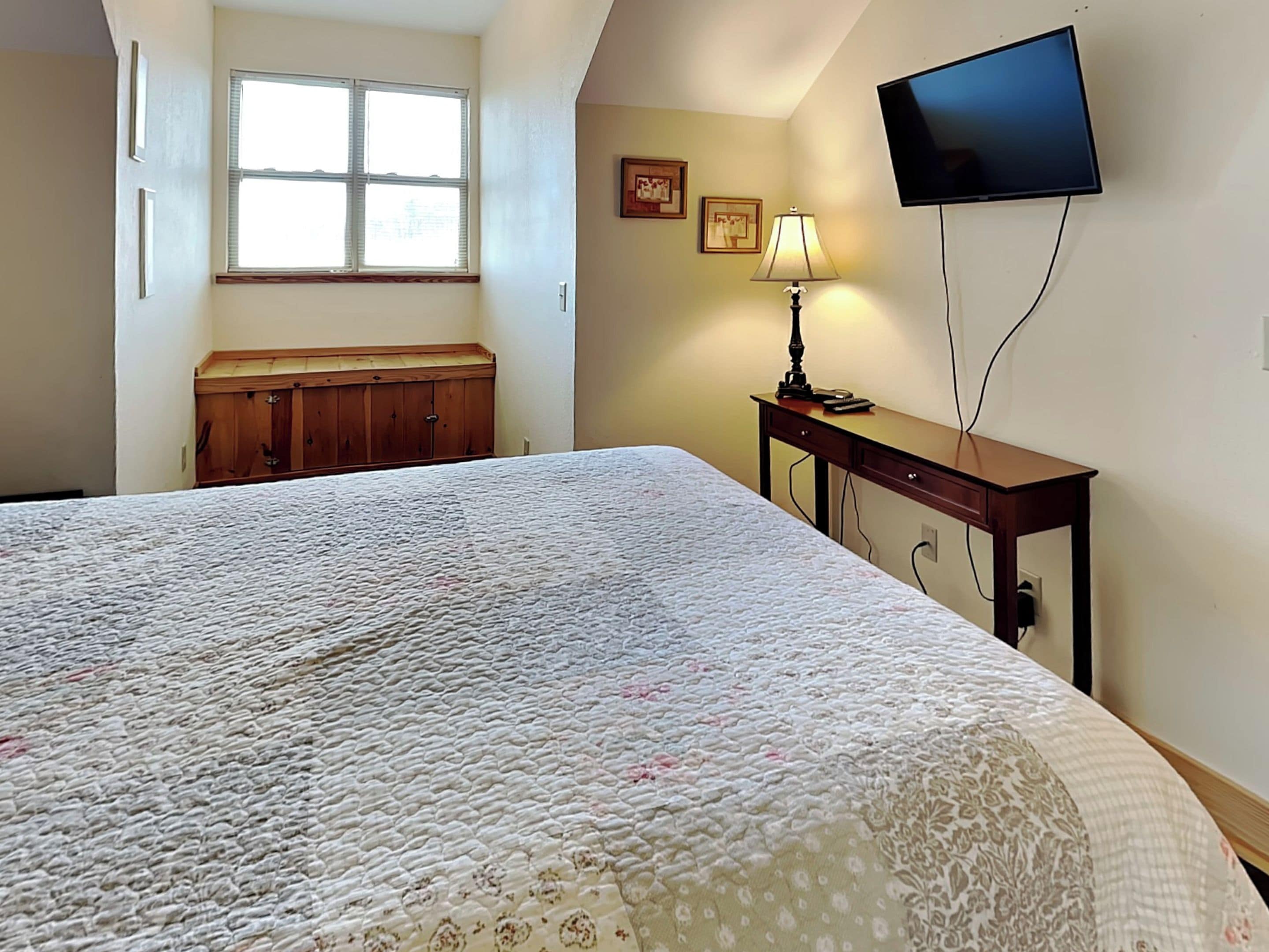 Ein Schlafzimmer mit einem großen Bett, einer Holzkommode, einem Schreibtisch mit Lampe und einem an der Wand befestigten Fernseher.