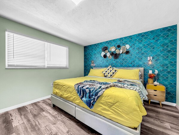 Un dormitorio con cama, mesita de noche, pared decorada con un motivo azul y ventana con persianas.