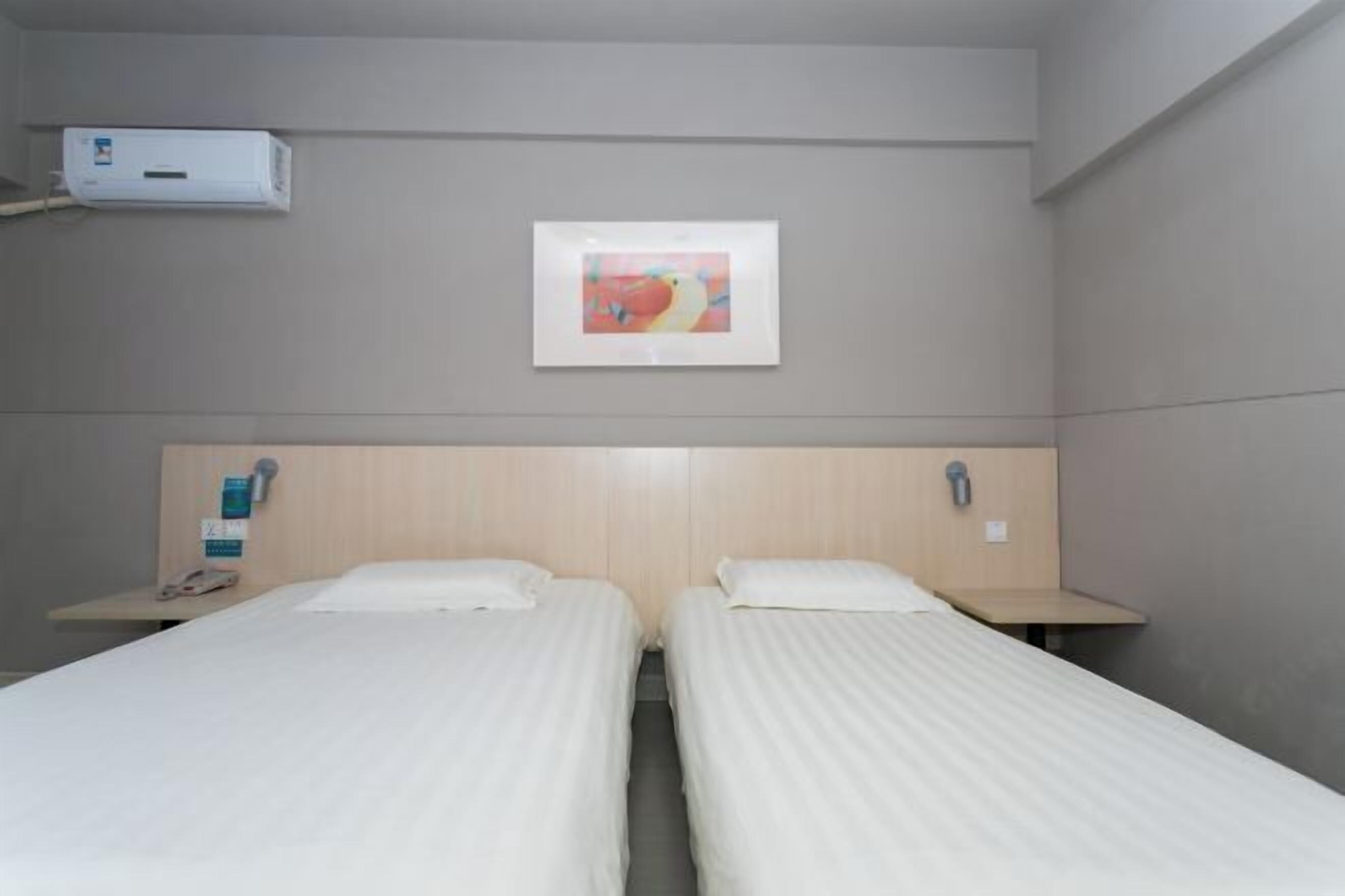 Habitación de hotel con dos camas individuales, una mesita de noche con un teléfono y un objeto pequeño encima, aire acondicionado de pared y un cuadro enmarcado sobre las camas.