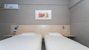 Habitación de hotel con dos camas individuales, una mesita de noche con un teléfono y un objeto pequeño encima, aire acondicionado de pared y un cuadro enmarcado sobre las camas.