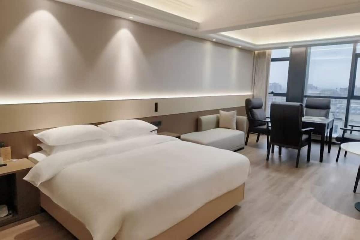 A hotel room with a large bed, a desk, and a chair. There is a view of the city through large windows.