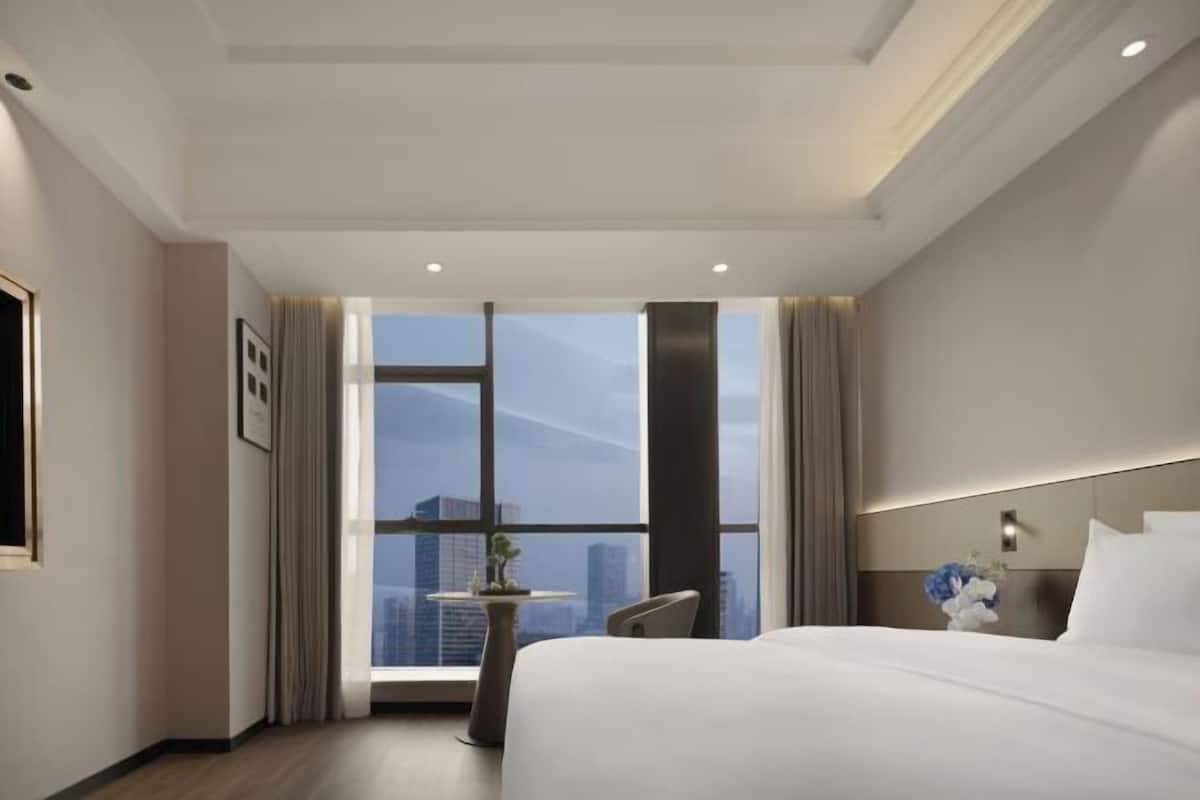 A hotel room with a large window, a bed, a small table with a plant, and a nightstand with a vase.