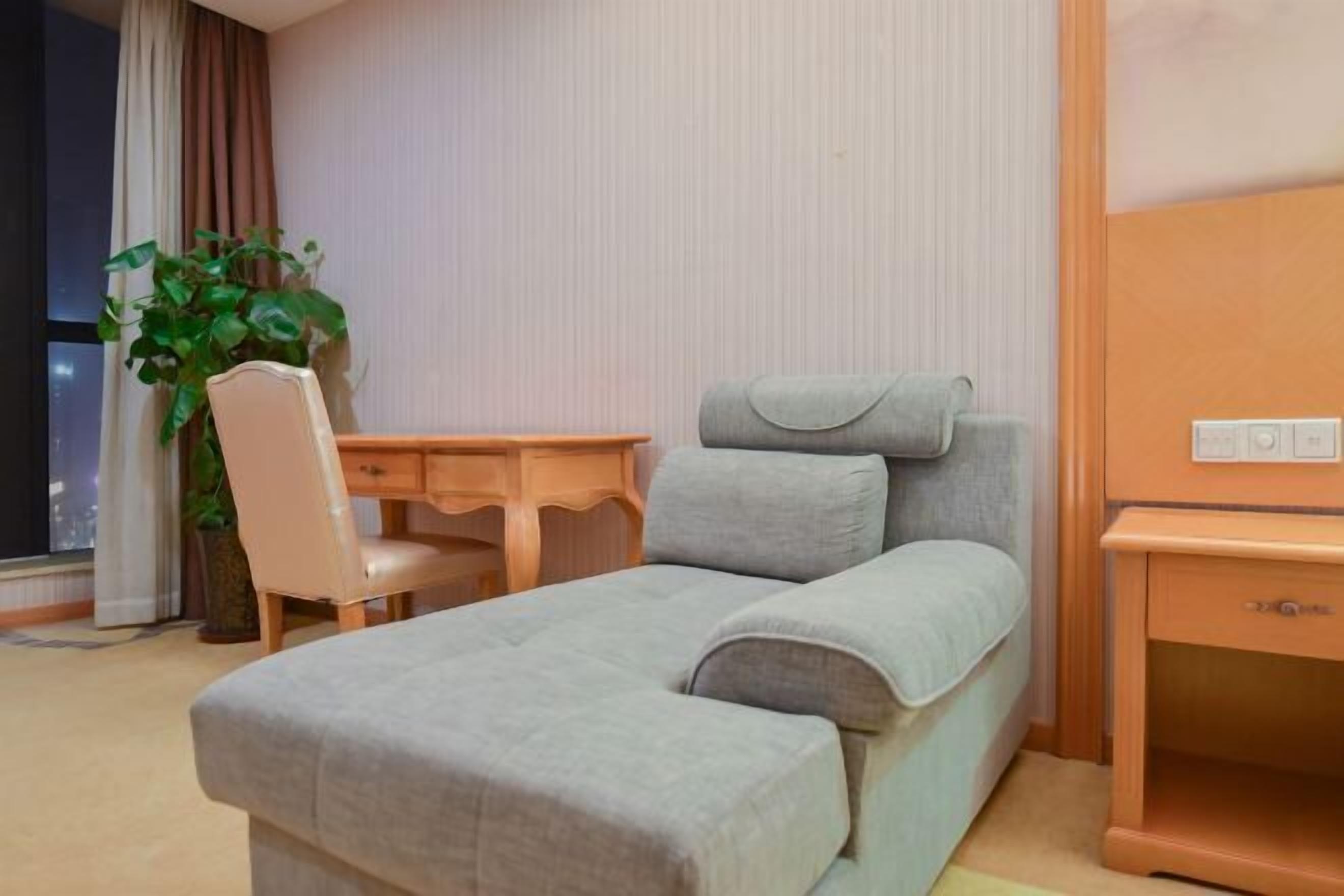 Una cama individual con colchón y almohadas grises, un escritorio de madera con una silla, una mesita de noche pequeña y una planta en maceta.