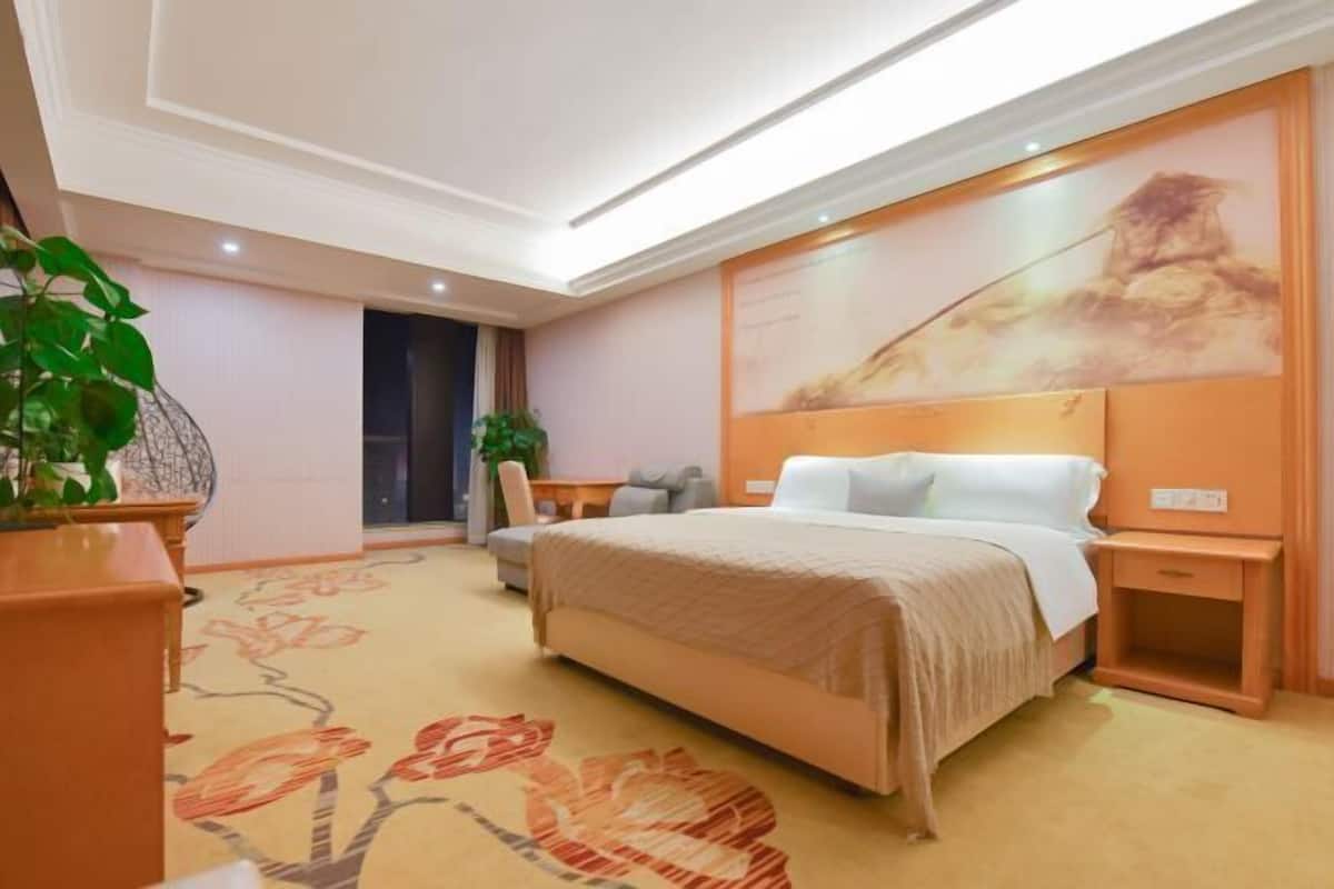 A hotel room with a large bed, a desk, a chair, and a potted plant.