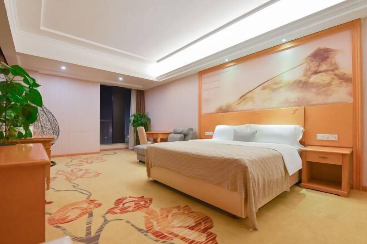 A hotel room with a large bed, a desk, a chair, and a potted plant.