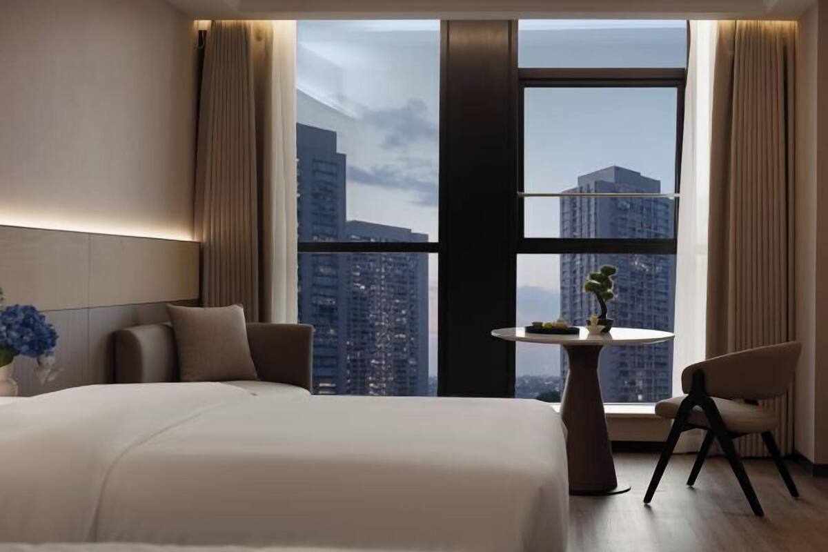 A hotel room with a large bed, a round table with a plant, a chair, and a view of a cityscape through large windows.