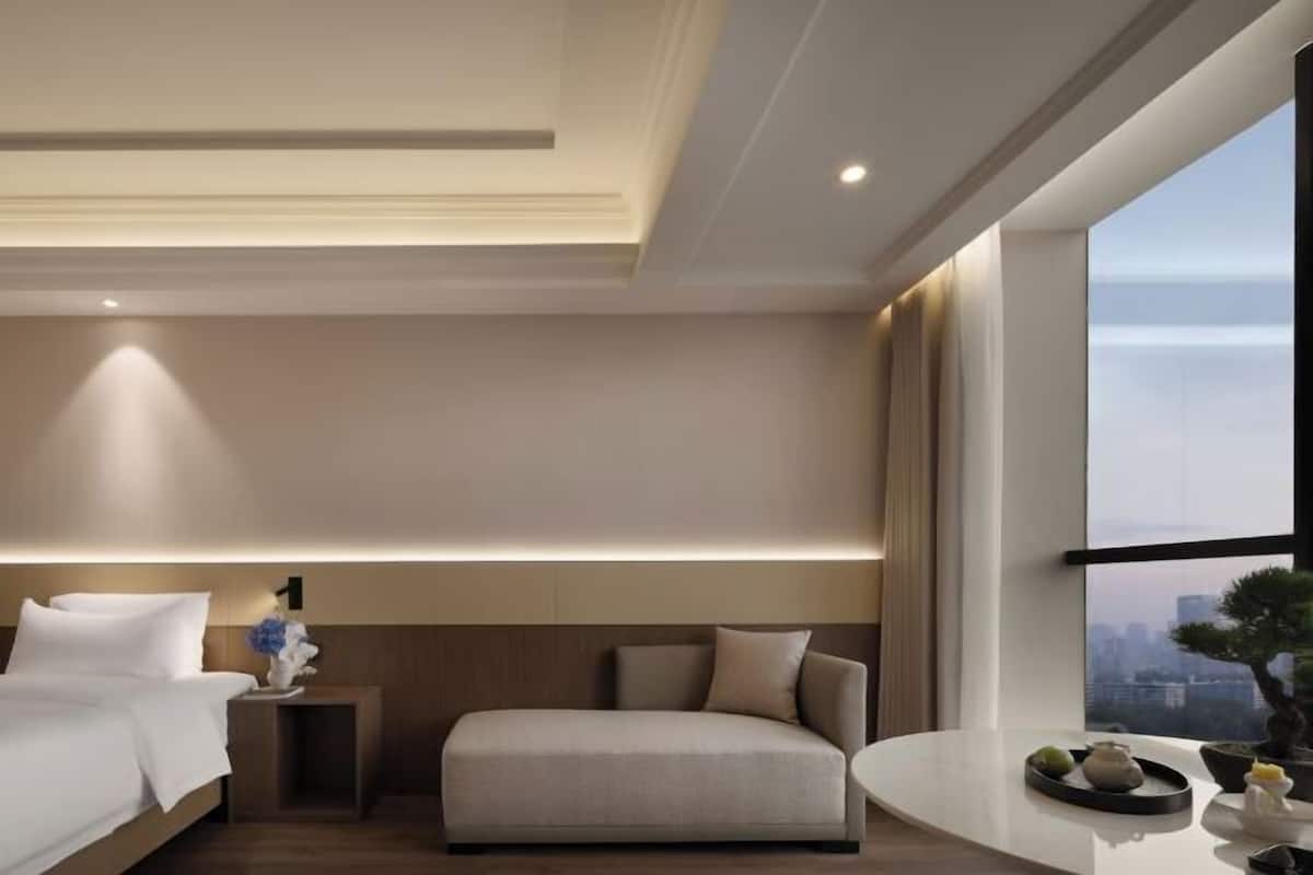 A modern hotel room with a bed, bedside table, and a view of the city through large windows.