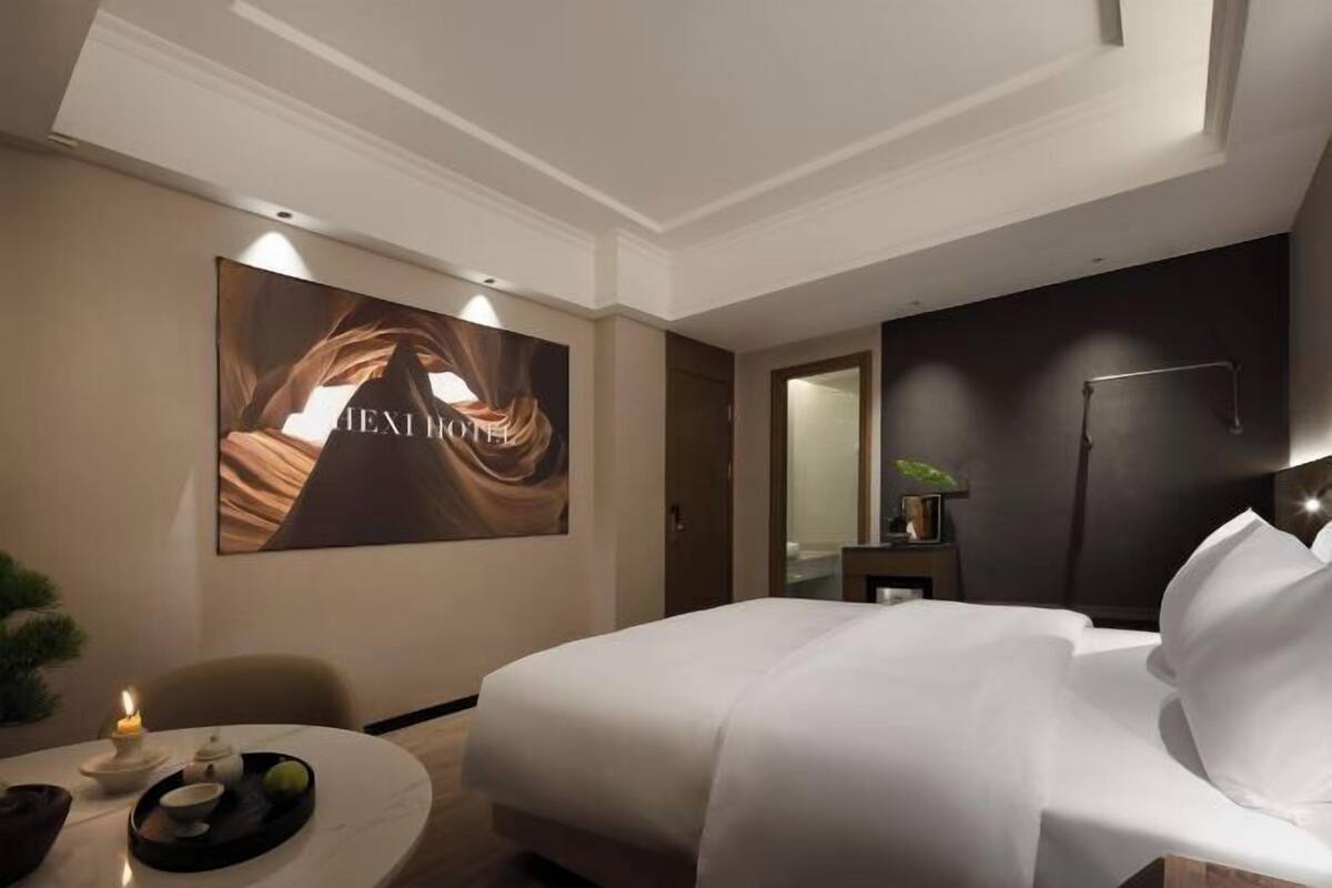 A hotel room with a large bed, a round table with a candle, a tray with a teapot and cups, a mirror, and a wall with a large framed picture.