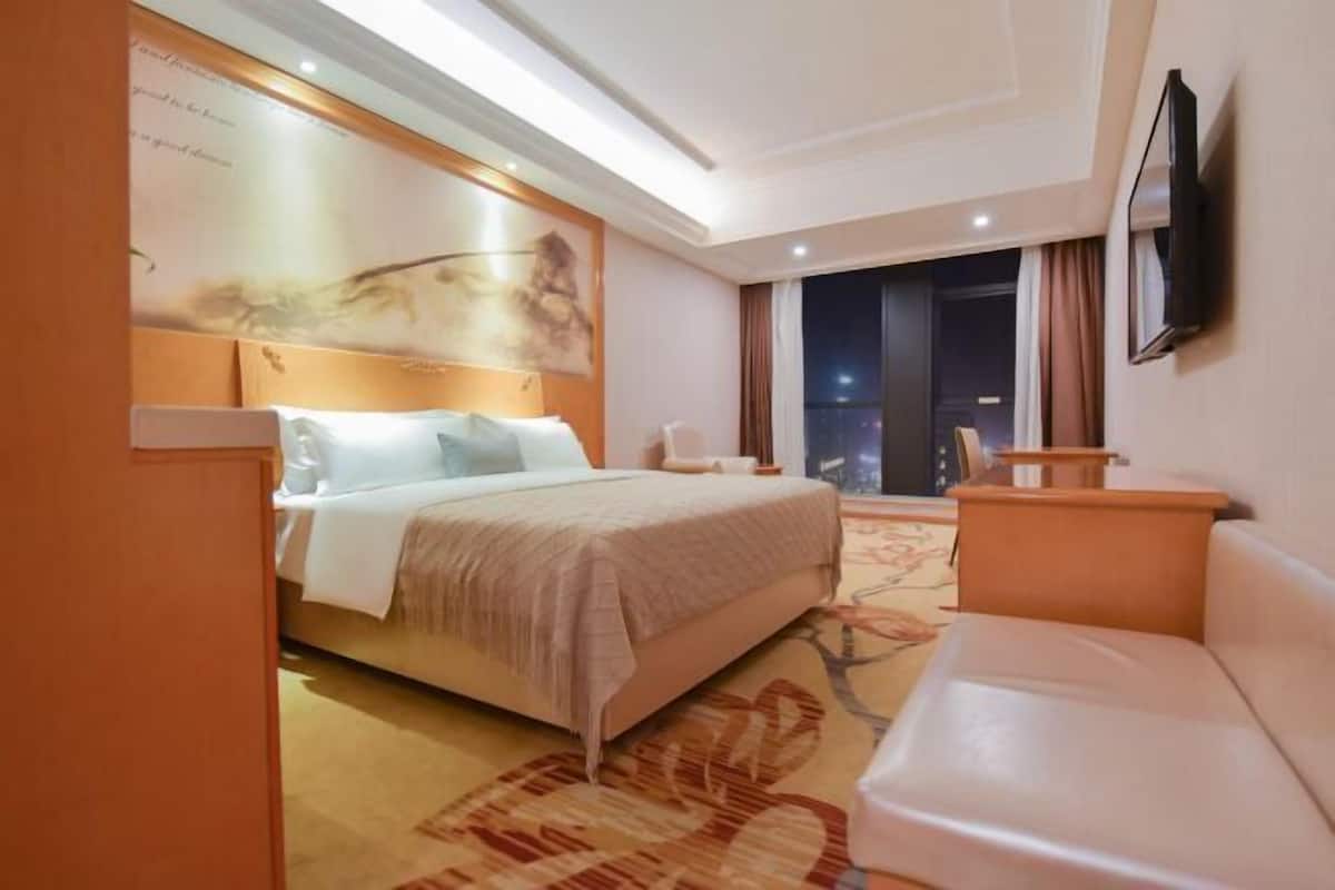 A hotel room with a bed, a sofa, a TV, and a large window with a view of the city at night.