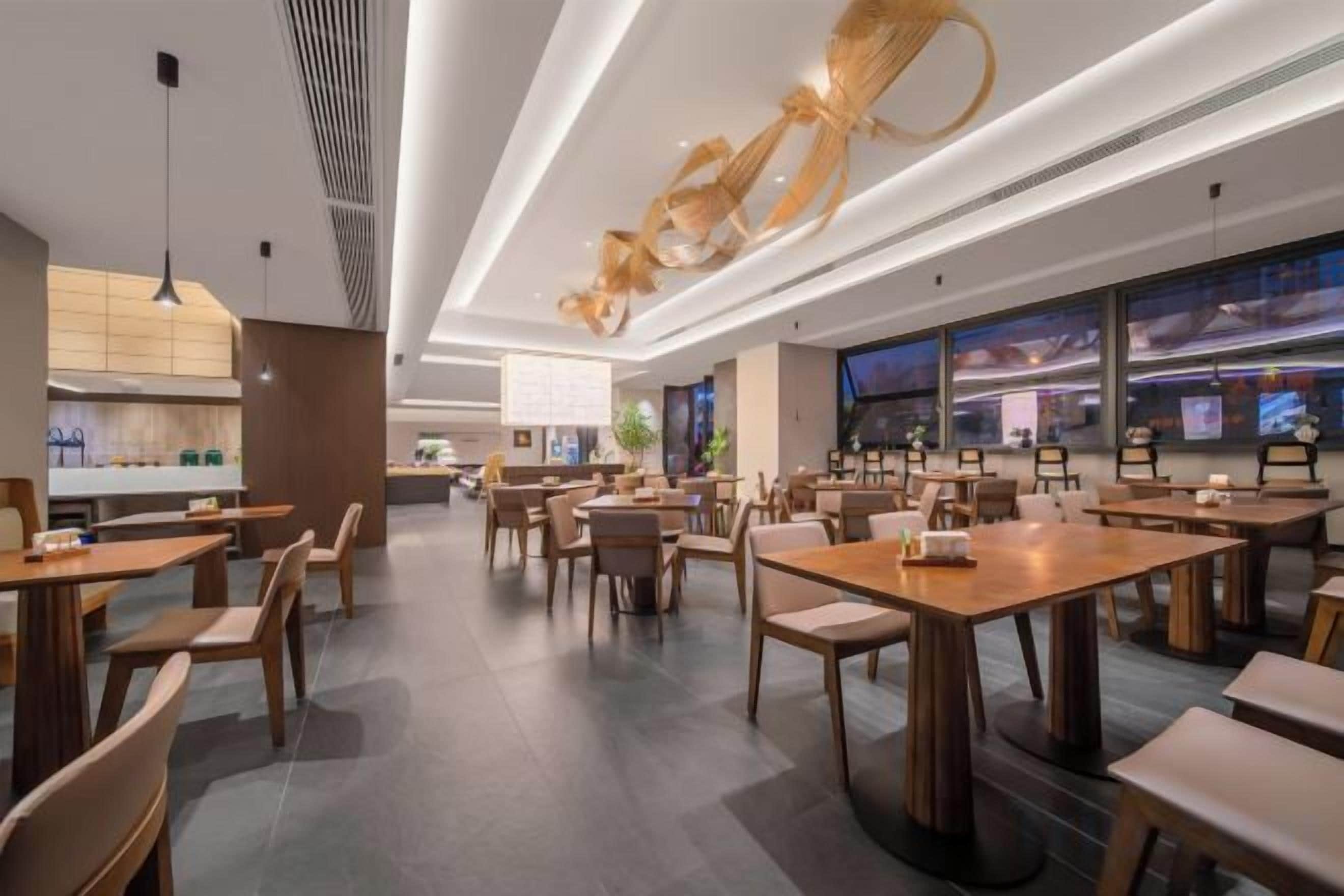 A modern restaurant interior with wooden tables and chairs, a kitchen area, and large windows.