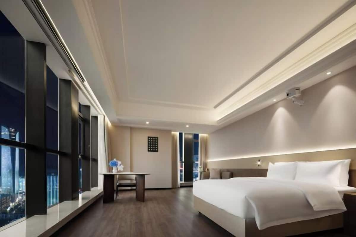 A modern hotel room with a large bed, a desk, and a view of the cityscape through floor-to-ceiling windows.