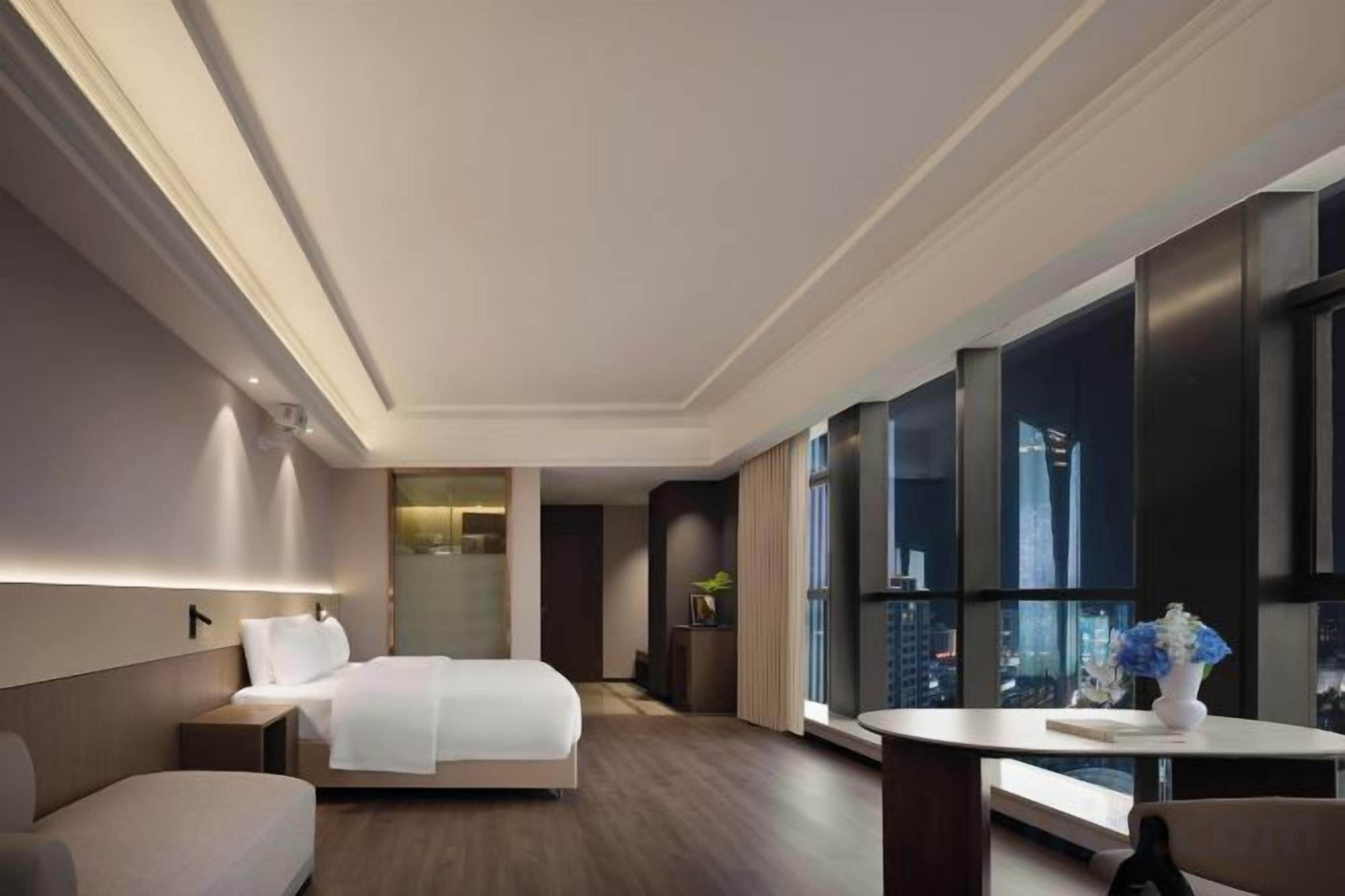 A modern hotel room with a large bed, a seating area, and a view of the cityscape through large windows.