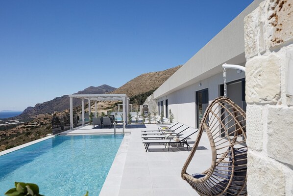 Ein moderner Poolbereich mit freiem Blick auf das Meer und die Berge, einer Pergola und Liegestühlen.