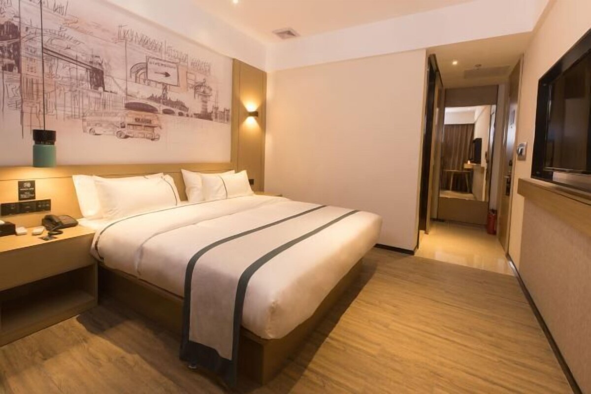 A hotel room with a large bed, a bedside table with a lamp, a wall-mounted artwork, and a door leading to another room.