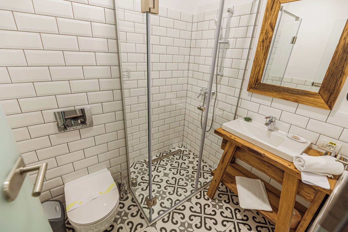 A bathroom with white tiled walls, a patterned floor, a shower with glass doors, a wooden vanity with a mirror, and a toilet.