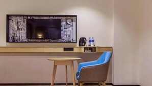 A modern room with a flat-screen TV mounted on the wall, a small wooden table, a blue and brown chair, and a shelf with a coffee maker and water bottles. - City Comfort Inn (Dongguan Gaobu Avenue) (Dongguan)