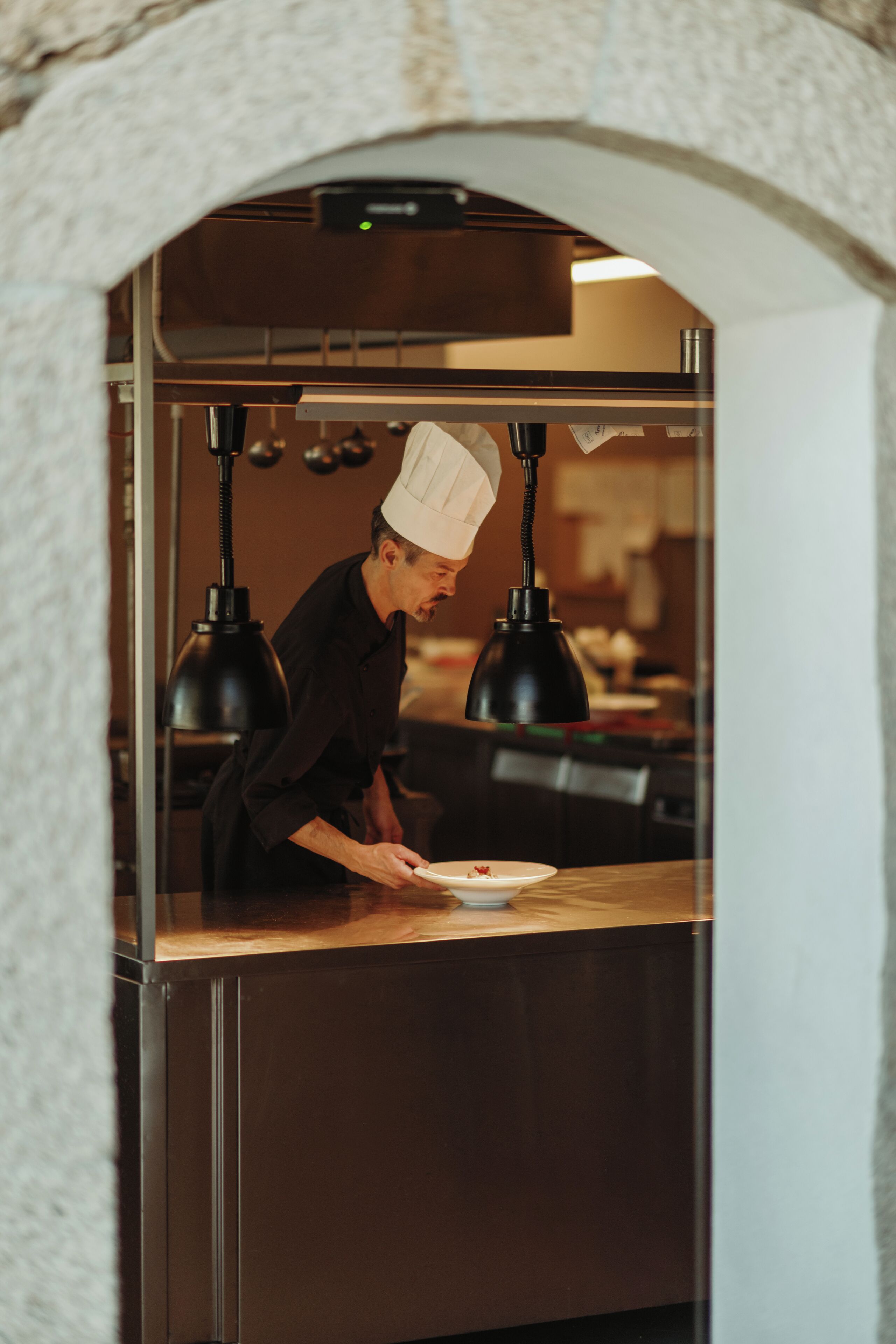 A chef in a professional kitchen with a stone archway, stainless steel appliances, and pendant lighting.