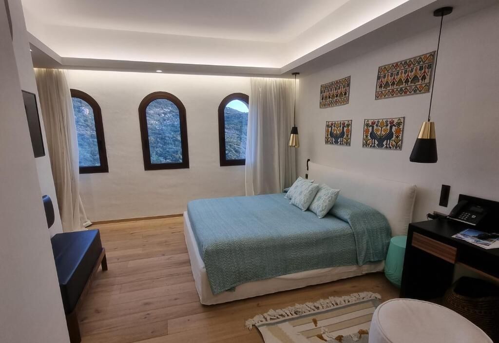 A bedroom with a bed, a desk, and a chair. There are three windows with curtains.