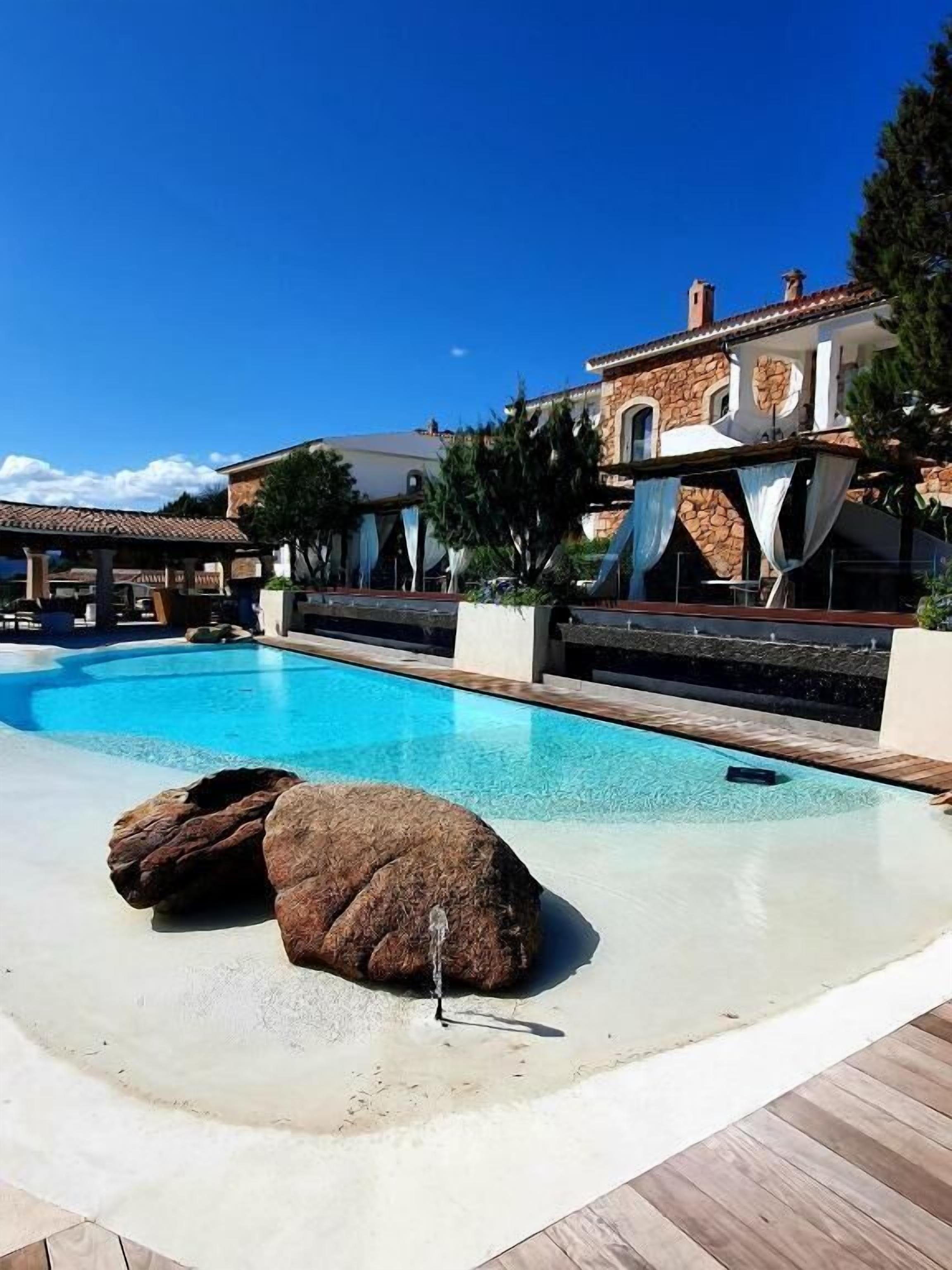A swimming pool with a rock feature, a covered area, and a building with stone and stucco exterior.