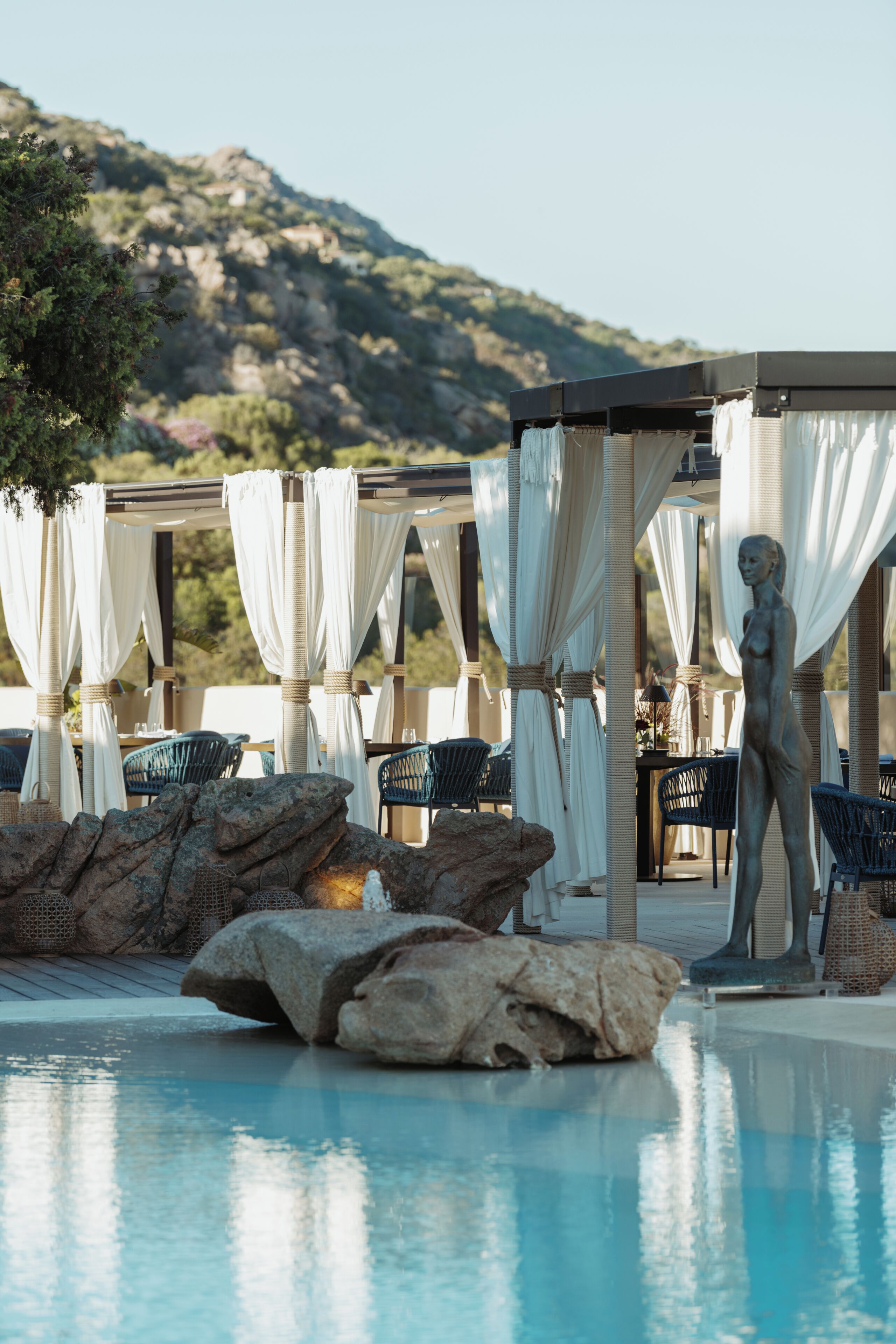 Zona de piscina amb gandules, una estàtua i un paisatge de muntanyes al fons.