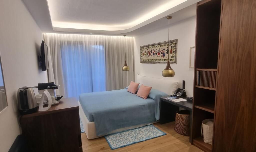 Una habitación de hotel moderna con cama, mesita de noche, lámpara y un ventanal con cortinas.