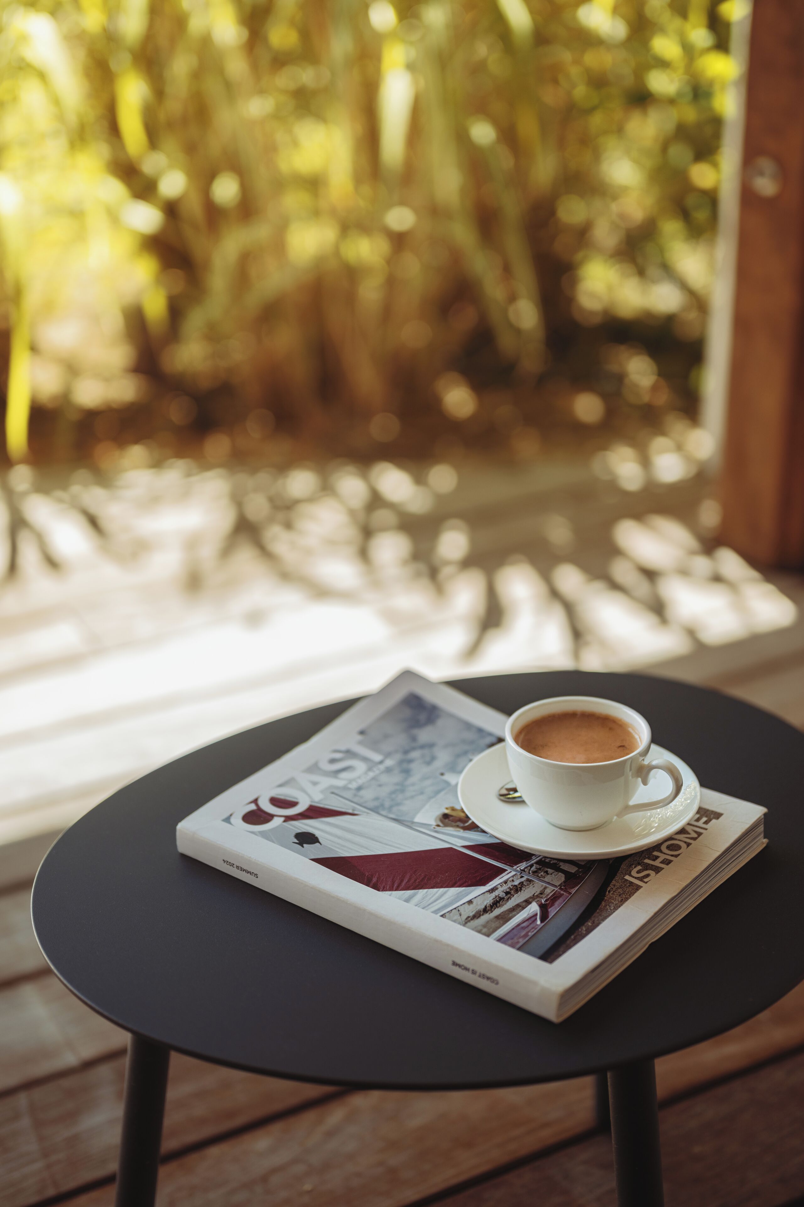 Une tasse de café sur une table, à côté d’un magazine, près d’une fenêtre donnant sur des arbres.