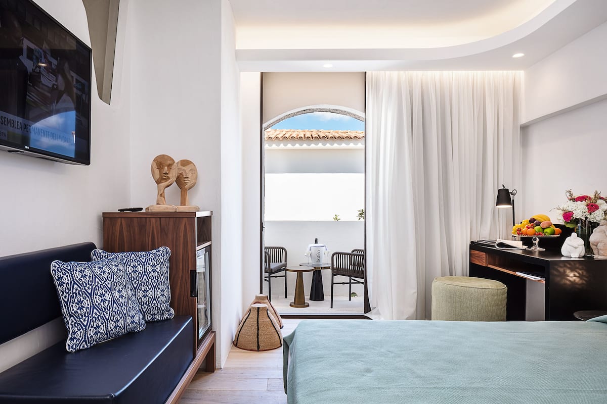 A modern hotel room with a bed, a sofa, a desk with a lamp, and a TV on the wall.
