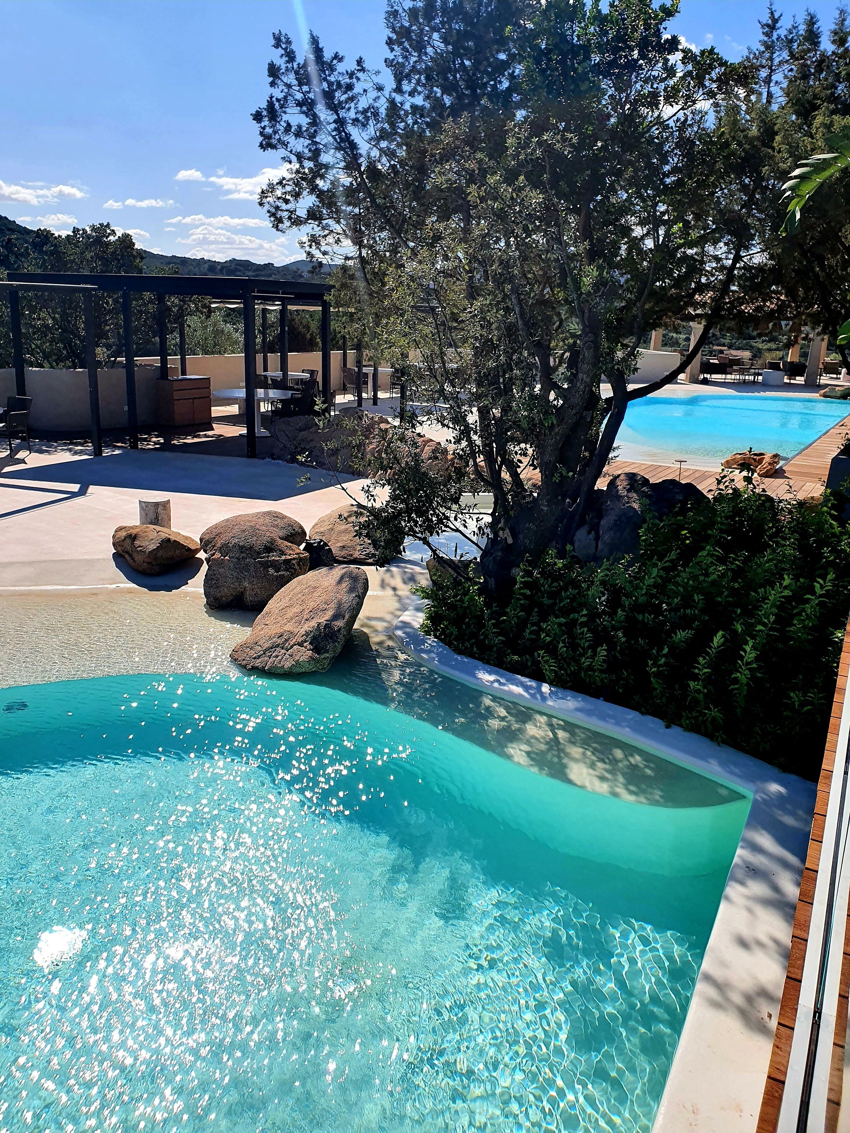 Ein klares Schwimmbecken mit Felsen, umgeben von viel Grün und einer Pergola.
