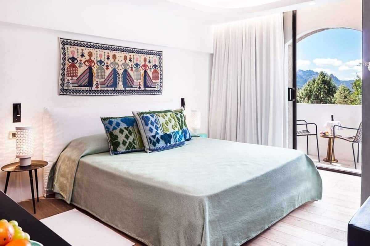 A bedroom with a bed, a nightstand, a wall-mounted lamp, a painting, and a balcony with a table and chairs.