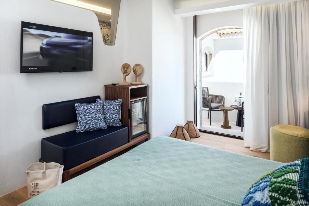 A hotel room with a bed, a TV mounted on the wall, a sofa with patterned cushions, and a view of the outdoor seating area through a glass door.