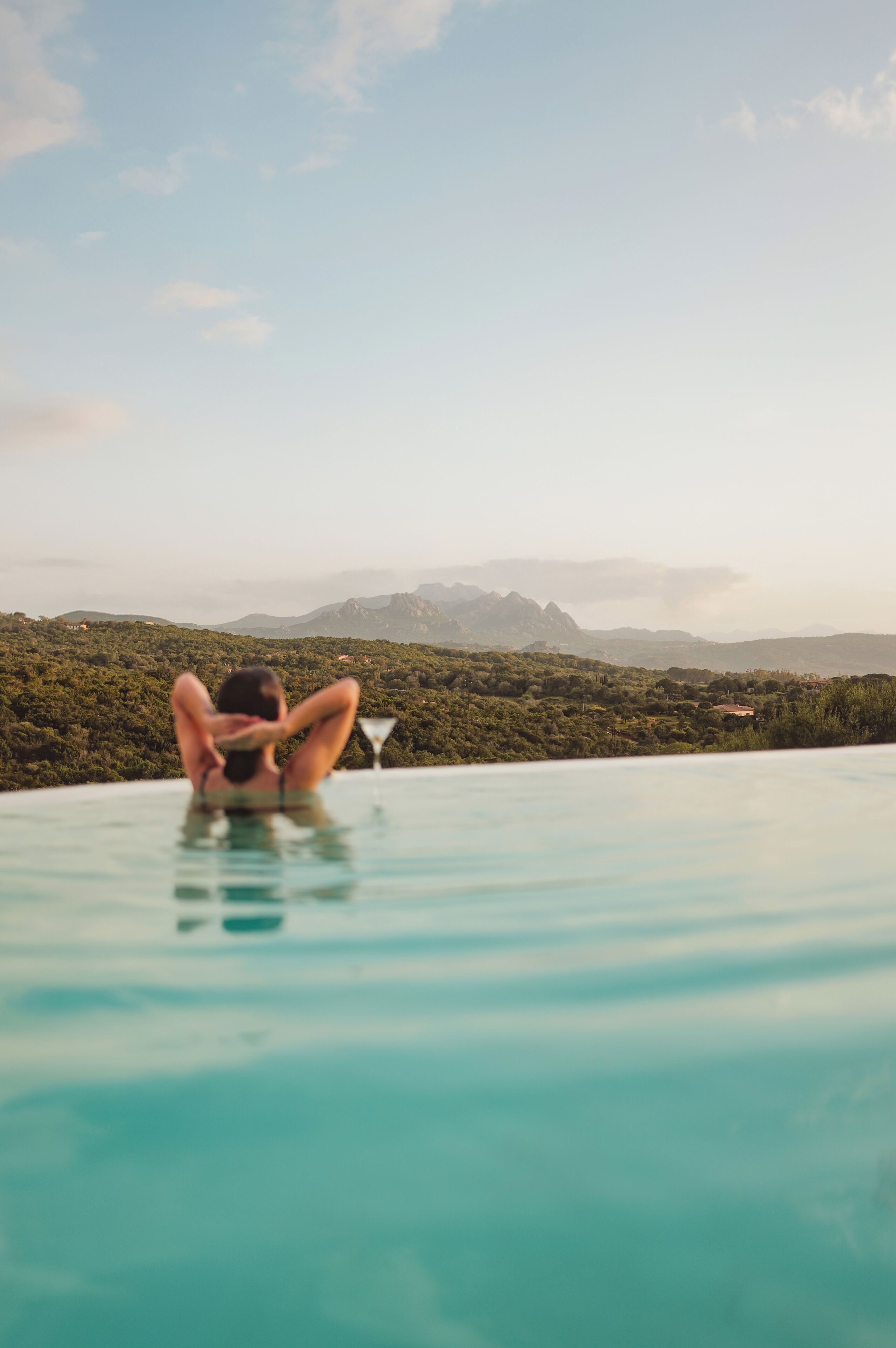 Eine Person entspannt in einem klaren, blauen Infinity-Pool mit Blick auf eine gebirgige Landschaft.