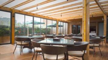 A covered outdoor dining area with round tables and wicker chairs, large windows, and a view of the outdoors.