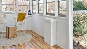 A room with a wooden floor, a hanging chair, a round rug, and large windows with a view of trees and a building. - Bohemian Styled Home with Bonus Game Room (Kannapolis)
