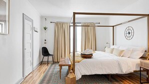 A four-poster bed with a white bedspread, a bedside table, a chair, and a patterned rug. - Bohemian Styled Home with Bonus Game Room (Kannapolis)