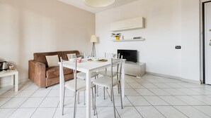 A modern living room with a brown sofa, white dining table, and chairs. There is a flat-screen TV on a stand, a lamp, and a small plant on a shelf. - HQ Aparthotel Milano Inn - Smart Suites -  Standard one bedroom Apartment (Cinisello Balsamo)