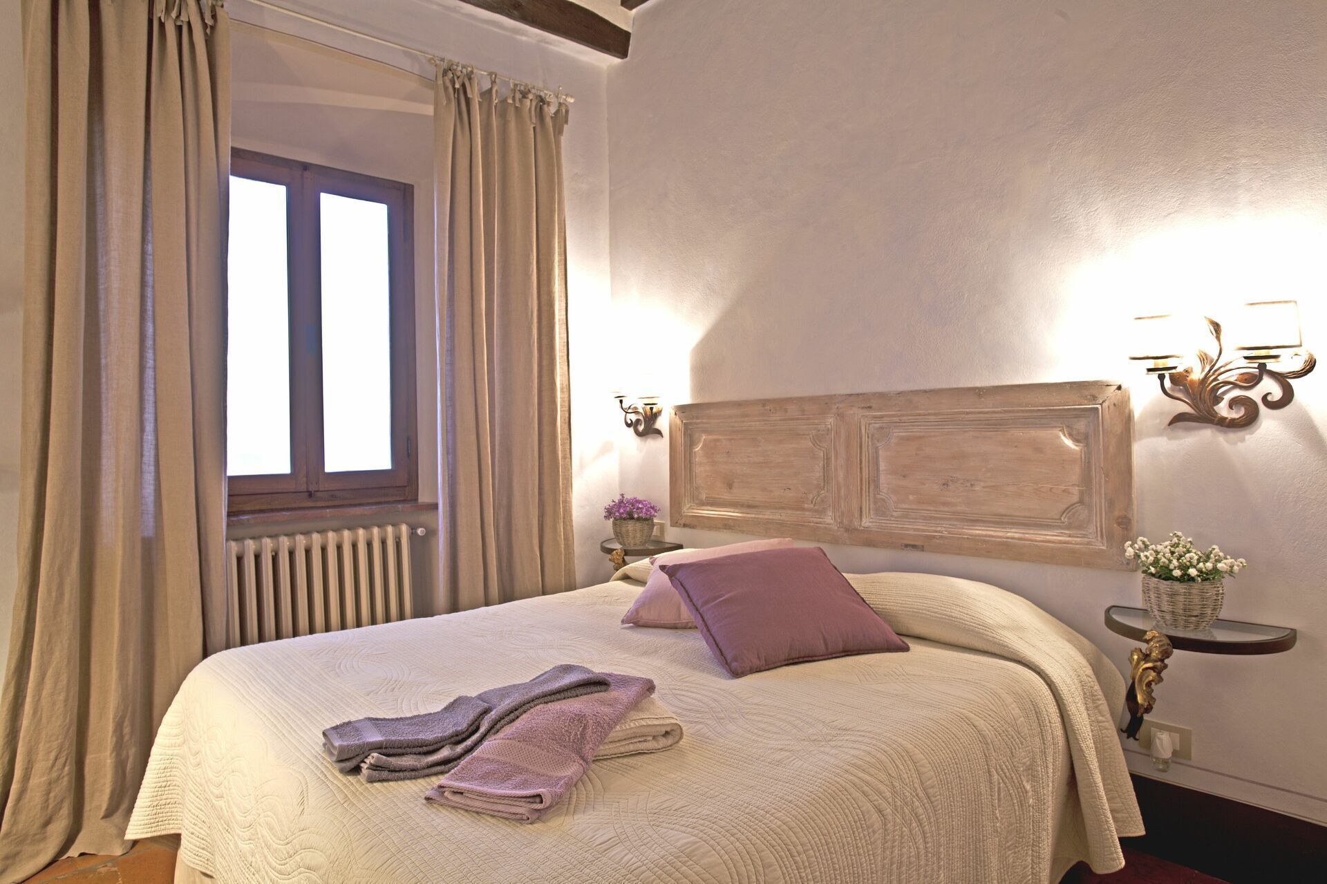 A neatly made bed with a headboard, a window with curtains, a wall-mounted light fixture, and a small bedside table with a vase of flowers.