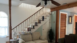Un salon avec un canapé beige, un canapé vert, un escalier en bois et un ventilateur de plafond.